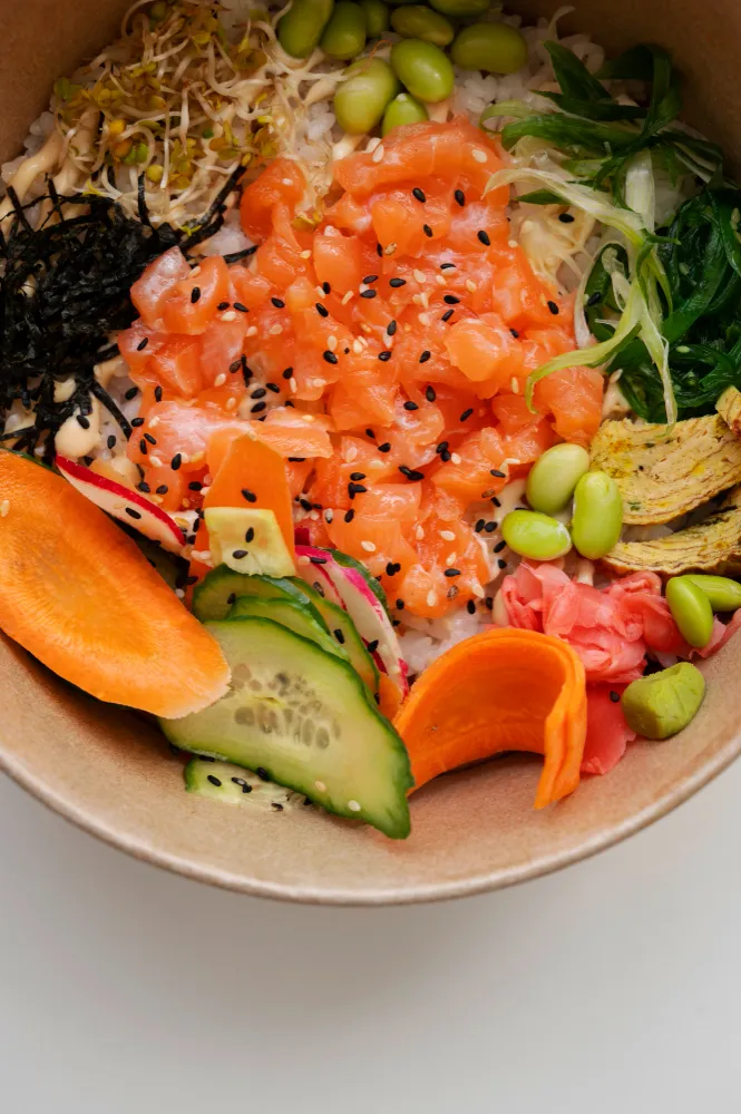 Salade au saumon cru sur riz, garni d'edamame, concombre, gingembre rose, algues wakame et nori, pousses et graines de sésame, servi dans un bol en carton kraft